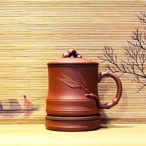 May include: A brown ceramic tea mug with a lid and a bamboo design. The mug has a handle and sits on a matching base.