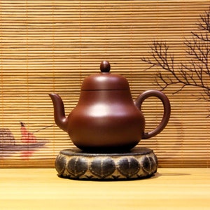 May include: A brown ceramic teapot with a rounded body and a lid, sitting on a black circular stand with a decorative design. The teapot is on a wooden surface with a bamboo mat background.