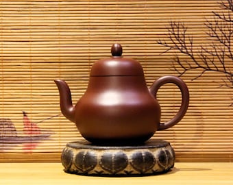 May include: A brown ceramic teapot with a rounded body and a lid, sitting on a black circular stand with a decorative design. The teapot is on a wooden surface with a bamboo mat background.