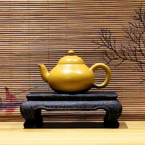 Op de afbeelding: Een mosterdgele keramische theepot met een afgeronde body, tuit en handvat, en een kleine knop op het deksel. De theepot staat op een donkere, sierlijke houten standaard. De achtergrond heeft een bamboe jaloezie en een decoratieve tak.