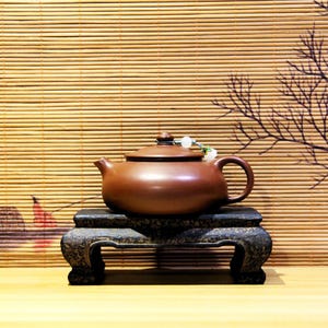 May include: A brown ceramic teapot with a lid and a handle, sitting on a dark wooden stand. The teapot has a string of white beads attached to the lid.