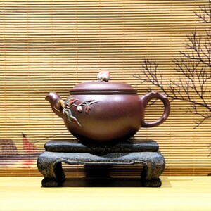 以下が含まれることがあります： 曲線的な取っ手と注ぎ口が付いた濃い茶色のティーポット。暗く装飾的な台の上に置かれています。ティーポットには花の装飾と蓋に小さな装飾的なつまみが付いています。背景には竹のブラインドと描かれた枝があります。