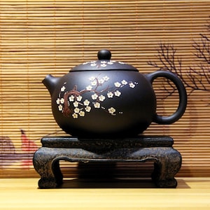 Könnte beinhalten: Schwarze Teekanne mit weißem Blumendesign auf einem schwarzen Holzständer. Die Teekanne hat einen runden Korpus und einen Ausguss an der Seite. Der Ständer hat drei Beine.