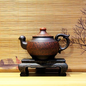 May include: A dark brown teapot with a red lid and a wooden-like pattern sits on a black wooden stand. The teapot has a curved spout and a handle.