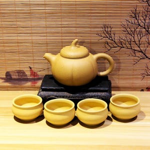 May include: A set of yellow ceramic teacups and teapot. The teapot has a rounded body and a lid with a small knob. The teacups are small and round. The set is arranged on a wooden surface.