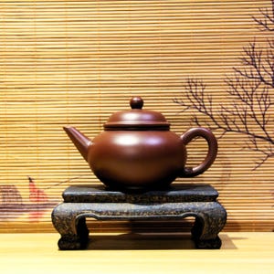以下が含まれることがあります： 黒い木製の台の上に、蓋つきの濃い茶色の急須が置かれています。急須は丸みを帯びたボディと側面に注ぎ口があります。台は3本の脚があり、木でできています。
