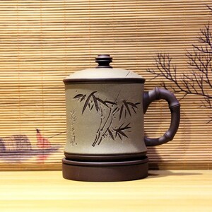 May include: A brown ceramic teacup with a lid and a handle. The cup has a design of bamboo plants and Chinese characters. The cup is sitting on a wooden surface.