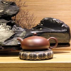 Può includere: Una teiera in ceramica marrone con un coperchio e un manico, seduta su una base con motivo bianco e nero. La teiera è davanti a una formazione rocciosa nera.
