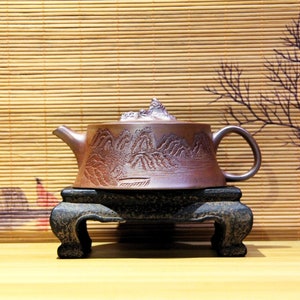 Puede incluir: Una tetera de cerámica marrón con un diseño de escena de montaña se asienta sobre un soporte de madera negro. La tetera tiene un cuerpo redondeado y un pico en el lado izquierdo. El soporte tiene tres patas.