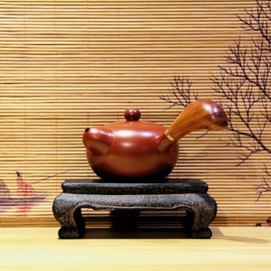 Può includere: Una teiera in terracotta rossa con un manico in legno si trova su un supporto in legno nero. La teiera ha un corpo arrotondato e un piccolo beccuccio. Il supporto ha un design semplice con tre gambe.