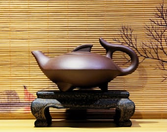 May include: A brown ceramic teapot with a unique fish-like design sits on a black wooden stand. The teapot has a spout and a handle, and the stand has three legs.