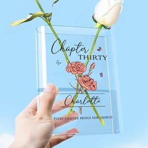May include: A clear glass vase with two white roses. The vase has the words "Chapter Thirty Charlotte" and a floral design. The text "EVERY CHAPTER BRINGS NEW GROWTH" is also on the vase. The vase is held against a blue sky.