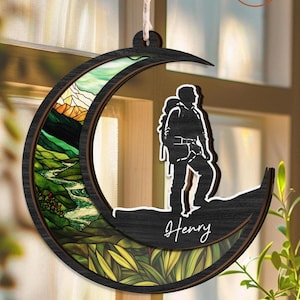 May include: A wooden moon-shaped ornament with a stained glass design of a mountain landscape. A silhouette of a person with a backpack is standing on a mountain peak. The ornament is personalized with the name "Henry".