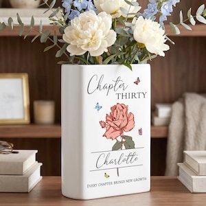 May include: A white rectangular ceramic vase featuring a printed design of a coral-colored rose, small butterflies, and the text "Chapter THIRTY", "Charlotte", and "EVERY CHAPTER BRINGS NEW GROWTH". The vase is filled with white peonies and blue delphiniums.