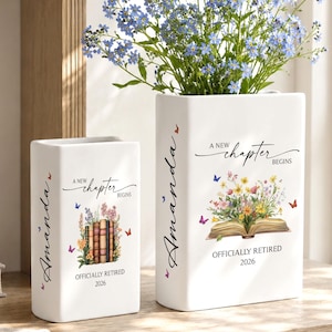 May include: Two white ceramic vases with floral designs and the text "A New Chapter Begins" and "Officially Retired 2026". One vase holds blue flowers. The vases are decorated with butterflies and the name "Amanda".