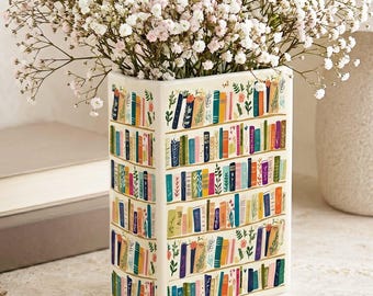 May include: A rectangular ceramic vase with a bookshelf design, filled with white and pink baby's breath flowers. The vase features colorful book spines and floral accents. The vase is approximately 8 inches tall.