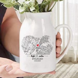 May include: White ceramic pitcher with a black map design and a red heart marking "Hell's Kitchen." The pitcher has a curved handle and is inscribed with "Kyle & Nadia ENGAGED 03.02.2025." The pitcher holds white flowers and greenery.