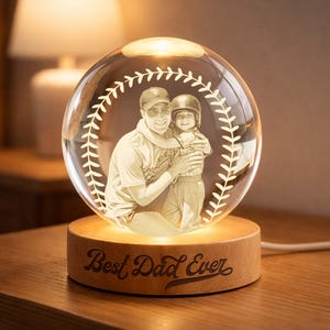 May include: A clear glass sphere with a baseball stitch design, featuring an engraved image of a father and child. The sphere sits on a wooden base with the text "Best Dad Ever". The base is illuminated, creating a warm glow.