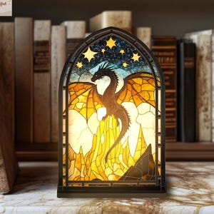 May include: A stained glass-style night light featuring a dragon design. The dragon is brown with orange wings, set against a backdrop of a starry night sky and yellow mountains. The night light has a black frame and is placed on a marble surface.