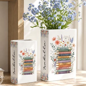 May include: Two white ceramic vases, one tall and one short, decorated with floral and book designs. The taller vase holds blue flowers. The word "Isabella" is written on the side of each vase. The vases are on a wooden surface.