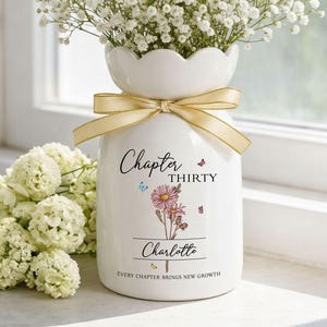 May include: White ceramic vase with a scalloped rim, tied with a gold ribbon, and filled with white flowers. The vase is decorated with the words "Chapter Thirty" and "Charlotte", along with a floral design and butterflies. The text "Every Chapter Brings New Growth" is also included.