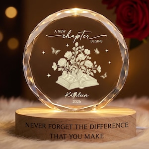 May include: A circular crystal plaque with faceted edges, resting on a light wood base. The crystal features an engraved open book with flowers, butterflies, and the text A NEW CHAPTER BEGINS, Kathleen, and 2026. The wood base reads NEVER FORGET THE DIFFERENCE THAT YOU MAKE.