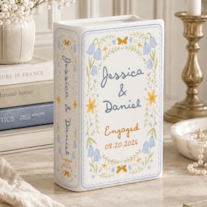 May include: A white ceramic book-shaped vase featuring blue and yellow floral illustrations, butterflies, and the text "Jessica & Daniel" with "Engaged" and "08.20.2026" printed on the front and spine in a decorative blue and orange script.