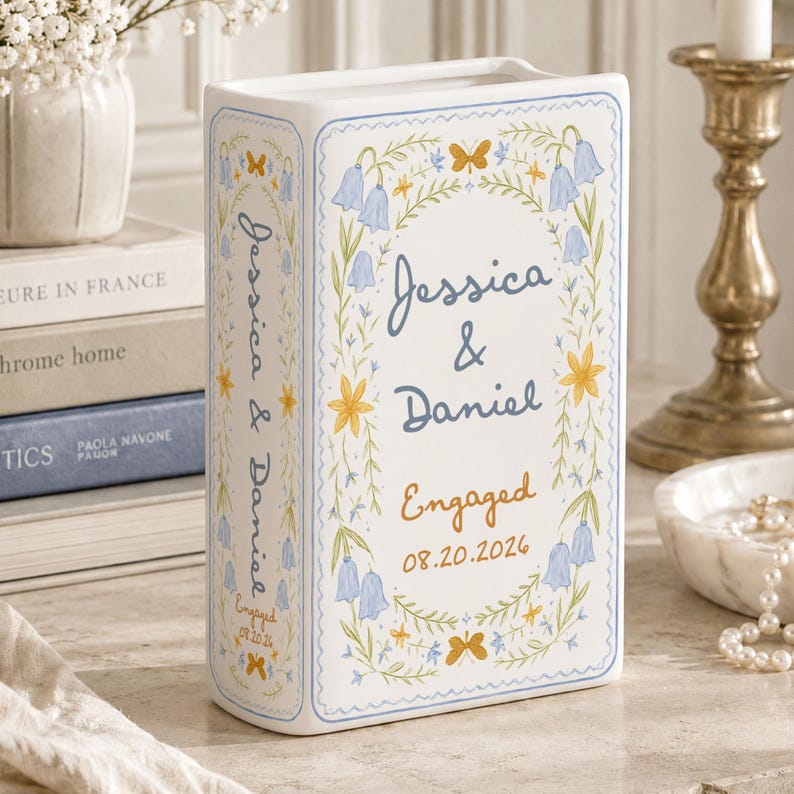 May include: A white ceramic book-shaped vase featuring blue and yellow floral illustrations, butterflies, and the text "Jessica & Daniel" with "Engaged" and "08.20.2026" printed on the front and spine in a decorative blue and orange script.