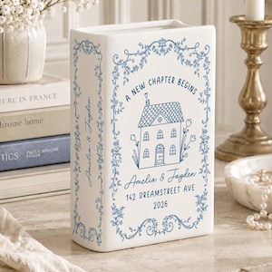May include: A white ceramic book-shaped vase featuring blue line art of a house, floral borders, and the text A NEW CHAPTER BEGINS, Amelia & Jayden, 142 DREAMSTREET AVE, 2026. The spine displays the names Amelia & Jayden in blue script.