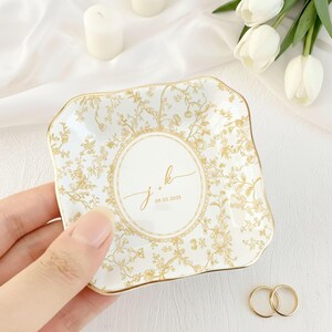 May include: A white and gold floral patterned ceramic ring dish with a scalloped edge. The dish features the initials "j + k" and the date "09.20.2025" in the center. Two gold wedding rings are next to the dish.