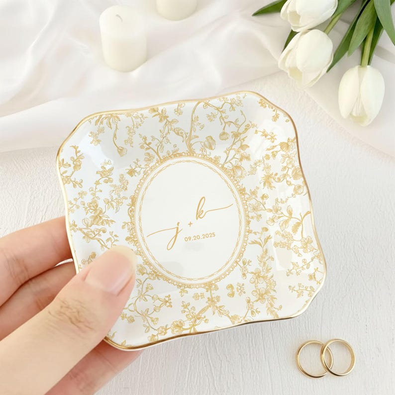 May include: A white and gold floral patterned ceramic ring dish with a scalloped edge. The dish features the initials "j + k" and the date "09.20.2025" in the center. Two gold wedding rings are next to the dish.