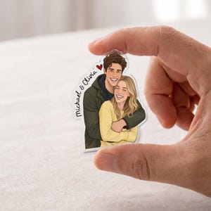 May include: A clear acrylic charm featuring a cartoon illustration of a smiling couple embracing. The man wears a dark green jacket, and the woman wears a yellow sweater. The charm has the names "Michael & Olivia" and a red heart.