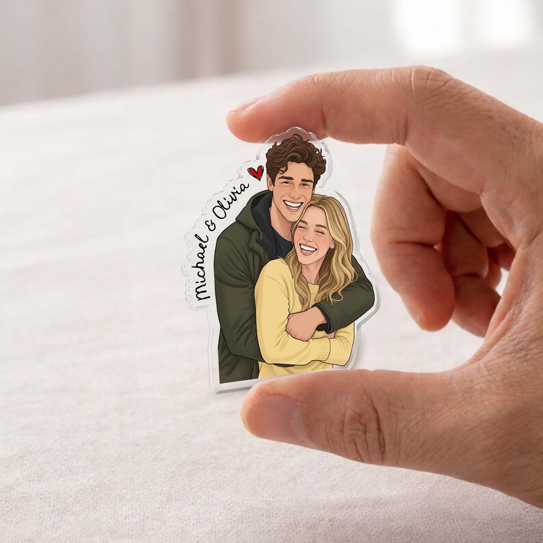 May include: A clear acrylic charm featuring a cartoon illustration of a smiling couple embracing. The man wears a dark green jacket, and the woman wears a yellow sweater. The charm has the names "Michael & Olivia" and a red heart.
