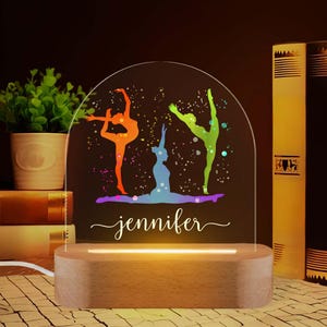 May include: A personalized night light featuring three colorful gymnast silhouettes in orange, green, and blue. The light has a wooden base and a clear acrylic top with the name "jennifer" in cursive.