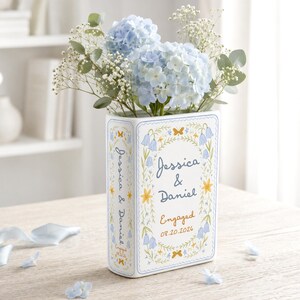 May include: A white ceramic book-shaped vase featuring blue and yellow floral illustrations, the text Jessica & Daniel, Engaged, and the date 08.20.2026. The vase is filled with light blue hydrangea flowers, baby's breath, and green eucalyptus leaves.