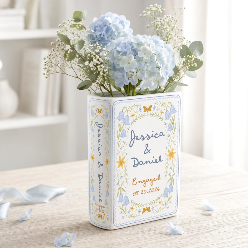 May include: A white ceramic book-shaped vase featuring blue and yellow floral illustrations, the text Jessica & Daniel, Engaged, and the date 08.20.2026. The vase is filled with light blue hydrangea flowers, baby's breath, and green eucalyptus leaves.