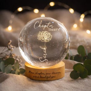 May include: A clear glass snow globe with the text "Chapter Thirty" and the name "Sarah" in script. A floral design is etched inside. The globe sits on a wooden base with the inscription "New chapter, new dreams, same beautiful you."