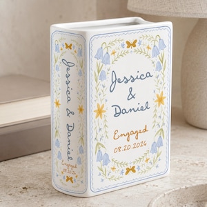 May include: A white ceramic book-shaped vase featuring blue and yellow floral illustrations, butterflies, and the text Jessica & Daniel Engaged 08.20.2026. The design includes a decorative border and is printed on both the front and the spine.