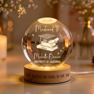 May include: A clear glass graduation sphere with the text "Mastered It!" and "2026". The sphere features a graduation cap, diploma, and books. The base is wood with the text "She believed she could, so she did".