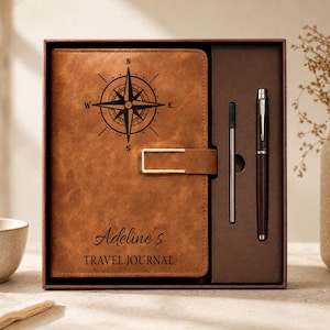 May include: A brown leather-like journal with a black compass graphic and the text Adeline's TRAVEL JOURNAL, featuring a gold-toned metal clasp. The journal is presented in a box alongside a matching brown pen and a thin black ink refill cartridge.
