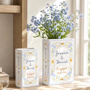 May include: Two rectangular white ceramic vases featuring blue and yellow floral illustrations, butterflies, and the text Jessica & Daniel Engaged 08.20.2026. The larger vase contains a bouquet of small blue forget-me-not flowers.