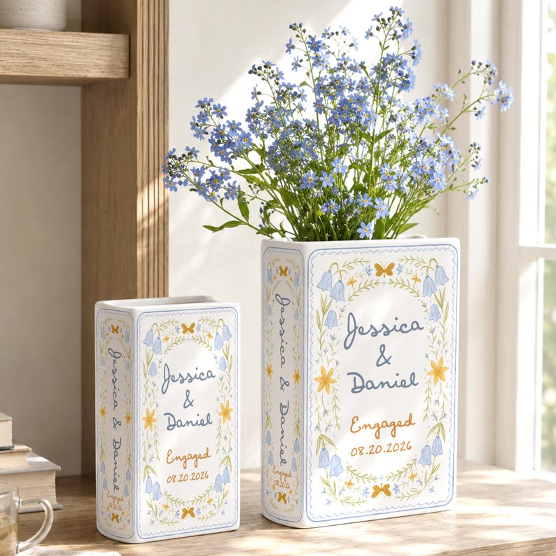 May include: Two rectangular white ceramic vases featuring blue and yellow floral illustrations, butterflies, and the text Jessica & Daniel Engaged 08.20.2026. The larger vase contains a bouquet of small blue forget-me-not flowers.