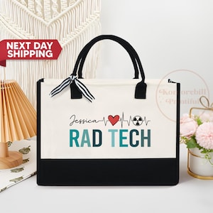 May include: A white canvas tote bag with black handles and a black bottom. The bag has a black and white ribbon tied around the handles. The bag has a red heart, a black radiation symbol, and the text "Jessica RAD TECH" printed on the front.