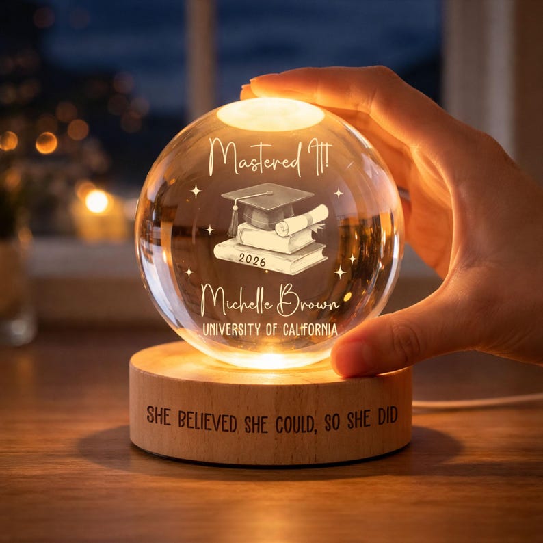 May include: A clear glass sphere with a graduation cap, books, and scroll design, illuminated from within. The sphere is engraved with the text "Mastered It!", "2026", "Michelle Brown", and "University of California". It sits on a wooden base with the inscription "SHE BELIEVED SHE COULD, SO SHE DID".