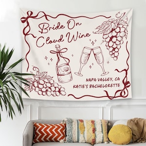 May include: Off-white wall tapestry with a burgundy border and illustrations of wine-related elements. The text "Bride On Cloud Wine" is displayed, along with a wine bottle, champagne glasses, and grapes. The text "Napa Valley, CA Katie's Bachelorette" is also included.