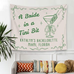 May include: A white tapestry with green text that reads "A Bride in a Tini Bit" and a green illustration of a martini glass with olives. The text below the illustration reads "KATHLYN'S BACHELORETTE MIAMI, FLORIDA."