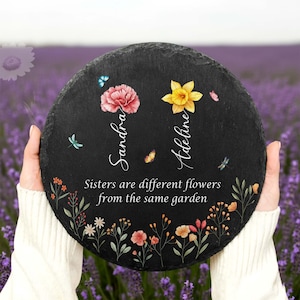 May include: A round, black slate plaque with floral and butterfly accents. The plaque features the names "Sandra" and "Adeline" with the quote "Sisters are different flowers from the same garden". The background is a field of purple flowers.