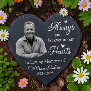 May include: Heart-shaped memorial stone with a black and white photo of a man. The inscription reads "Always and Forever in our Hearts" and "In Loving Memory of William Andrew 1952-2025". The stone is surrounded by flowers.