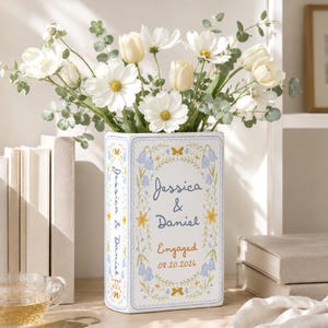 May include: A rectangular white ceramic vase featuring a blue and yellow floral border with the text Jessica & Daniel, Engaged, and 08.20.2026. The vase is filled with a bouquet of white cosmos flowers, cream tulips, and green eucalyptus stems.