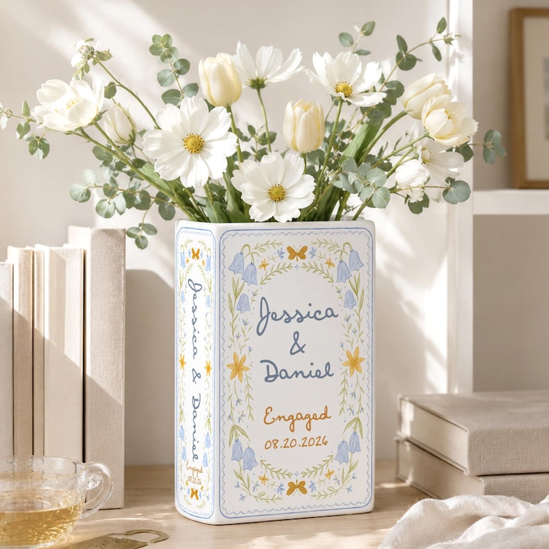 May include: A rectangular white ceramic vase featuring a blue and yellow floral border with the text Jessica & Daniel, Engaged, and 08.20.2026. The vase is filled with a bouquet of white cosmos flowers, cream tulips, and green eucalyptus stems.